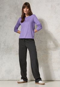 Hellpurpurfarbenes Langarmoberteil mit seitlichen Nähten, kombiniert mit locker sitzenden, dunkelgrauen Hosen, getragen mit weißen Sneakers vor einem neutralen Hintergrund.