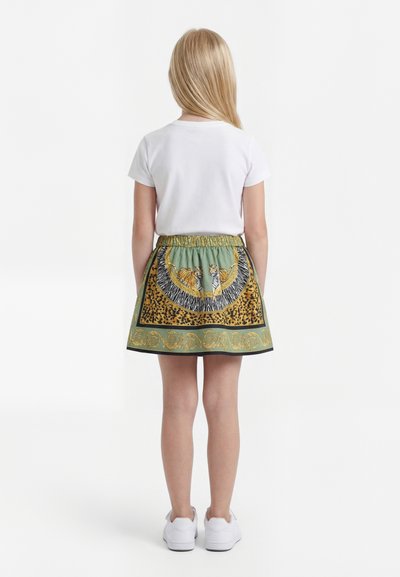 Kind met lang blond haar, draagt een wit t-shirt, groene rok met tijger- en luipaardprint en witte sneakers, staat met de rug naar voren.