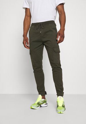Man draagt olijfgroene cargojoggers met verstelbare taille en neon groene hardloopschoenen, staand tegen een effen achtergrond.