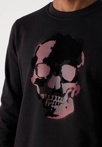 Hanorac negru cu un grafic mare, stilizat al unui skull în nuanțe de roz și negru, cu o textură netedă și guler rotund.