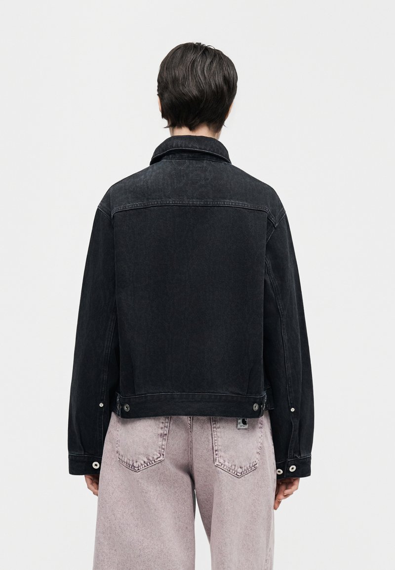 Persona con capelli corti e scuri che indossa una giacca di jeans nera e jeans viola chiaro, rivolta di spalle contro uno sfondo bianco semplice.