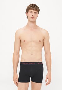 Schwarze Boxershorts mit einem roten Logo-Bund, aus einem glatten Stoff gefertigt und eng anliegend, von einer Person ohne Shirt präsentiert.