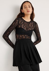 Μαύρο τοπ με μακριά μανίκια από δαντέλα με διάφανο σχέδιο πάνω σε μαύρο αμάνικο κορμάκι, συνδυασμένο με ψηλόμεση μαύρη φούστα μίνι με πιέτες.