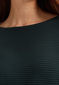 Top verde a coste con scollo ampio e tessuto testurizzato visibile. Scatto ravvicinato che mostra il motivo intricato e la superficie liscia.