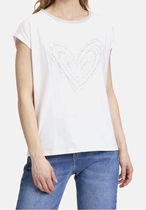 Vrouw draagt een wit kortärmelig shirt met een subtiel hartmotief van strassteentjes en blauwe spijkerbroek, tegen een effen achtergrond.