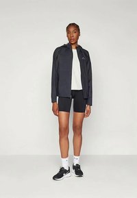 Černá lehká bunda s kapucí, v kombinaci s bílou košilí a černými šortkami. Oblečené s černými teniskami a bílými ponožkami, stojící na neutráln�ím pozadí.