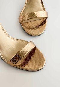 Arany szandálok texturált, kígyóbőr mintával. Széles pánt található a lábujjaknál, és sima belső bélés. Lapos talp.
