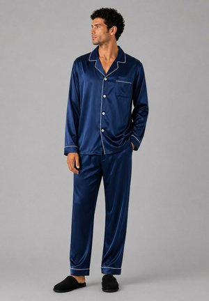Homme debout en pyjama satin bleu marine avec passepoil blanc, portant des chaussons noirs, regardant sur le côté sur fond gris.