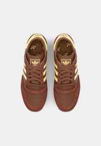 Brune sportsko med gule striper; laget av semsket skinn og tekstil. Har en rund tå og snøring; Adidas-logo på tungen.