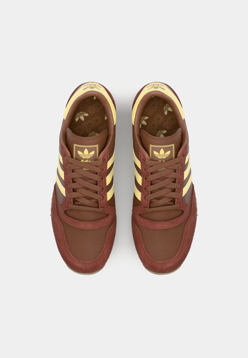 Brune sportsko med gule stribe accenter; lavet af ruskind og tekstil. Har en rund tå og snørebånd; Adidas-logo på tungen.
