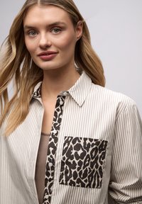 Chemise à rayures crème et beige avec une poche à imprimé léopard. Dotée d'un col classique et d'une texture de tissu douce.