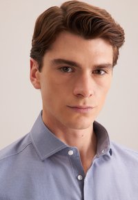 lichtblauwe blouse met een gestructureerd patroon, voorzien van een spread collar en witte knopen. Close-up van de bovenkant van de blouse.