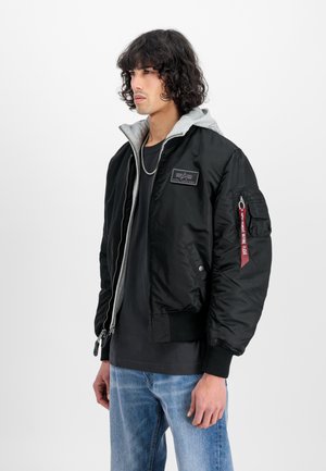 Giacca bomber nera con cappuccio grigio, chiusura con zip frontale, polsini a coste e tasca sulla spalla. Presenta una toppa con logo e un'etichetta di stoffa rossa sulla manica.