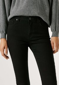 Jeans de mezclilla negra con un diseño ajustado, que cuentan con un estilo clásico de cinco bolsillos y costuras sutiles. Llevados con un suéter gris de canalé.