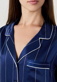 Mörkblå satinpyjamasöverdel med vita ränder, som har en notched krage, knappstängning och en enda ficka med kontrasterande kant.