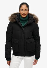 Sort pufferjakke med en hætte i faux fur, to frontlommer og ribbede manchetter. Båret over en teal strikket turtleneck.