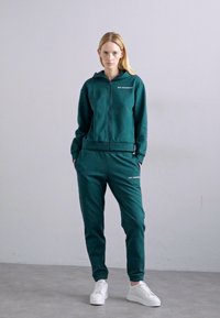 Γυναίκα στέκεται μπροστά σε έναν απλό τοίχο, φορώντας πράσινη φόρμα με φερμουάρ και αντίστοιχες φόρμες, με άσπρα αθλητικά παπούτσια, με τα χέρια στις τσέπες.