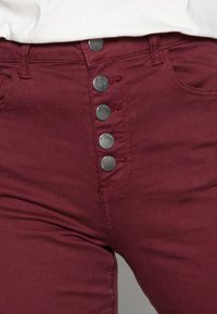 Jean taille haute bordeaux avec une fermeture à boutons sur le devant dotée de cinq boutons en métal, des poches latérales et une texture de tissu lisse et extensible.