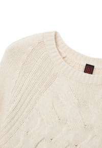Pull en maille épaisse crème avec motifs torsadés et côtes, avec un col rond côtelé et une petite étiquette en tissu noire et rouge à l'intérieur du col.