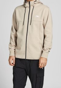 Felpa beige con zip e cappuccio regolabile, zip nera a contrasto e un piccolo logo sul petto. Presenta maniche lunghe e una vestibilità comoda.