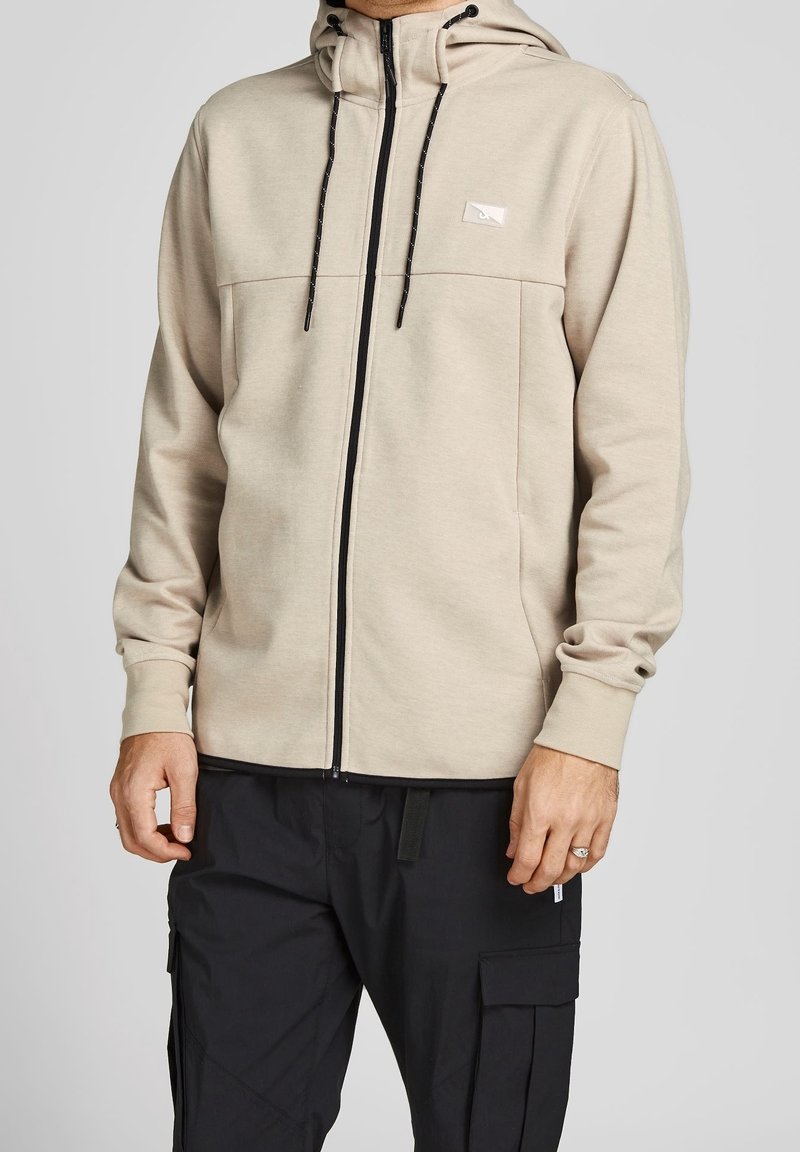 Felpa beige con zip e cappuccio regolabile, zip nera a contrasto e un piccolo logo sul petto. Presenta maniche lunghe e una vestibilità comoda.