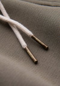 Deux cordons beiges de sweat à capuche avec embouts en métal bronze étiquetés "ombre" reposant sur un tissu taupe texturé.