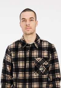 Camisa de franela a cuadros en negro y beige con cuello clásico, botones a presión, un bolsillo en el pecho y un parche de logo en el bolsillo.