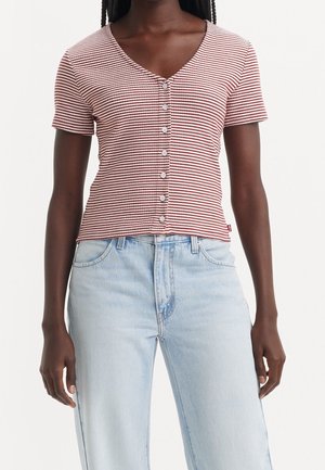 Femmes portant un haut à manches courtes, rayé rouge et blanc, boutonné, avec un jean taille haute bleu clair sur un fond uni.
