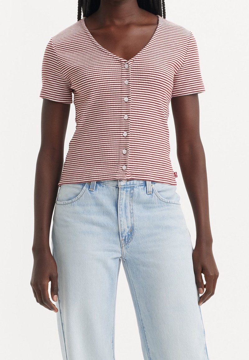 Femmes portant un haut à manches courtes, rayé rouge et blanc, boutonné, avec un jean taille haute bleu clair sur un fond uni.