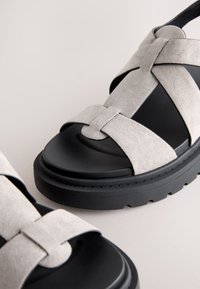 Sandales à semelle noire épaisse avec des brides croisées en daim gris clair et orteils ouverts sur un fond neutre.
