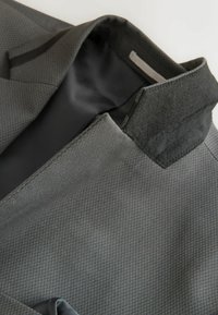 Blazer gris con un acabado texturizado, forro de satén negro y cuello de tela contrastante. Los detalles notables incluyen costuras y diseño de bolsillos.