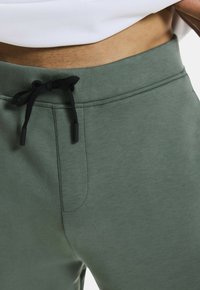 Lichtgroene joggingbroek van zacht materiaal, met een zwarte trekkoordsluiting, een zijzak en een gladde textuur.