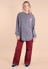 Femme se tenant debout portant une chemise à boutons à manches longues à motifs, un pantalon large bourgogne et des baskets noires sur un fond beige.