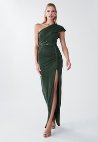 Žena v tmavozelených zamatových šatách s jedným ramenom, opaskom a vysokým rozparkom, doplnených o telové sandále na podpätku a zlaté náušnice.
