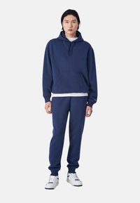 Navyblauer Hoodie und passende Sweatpants aus weichem Material. Mit einer Kängurutasche, ribbed Bündchen und einem darunterliegenden, mehrlagigen weißen Shirt.