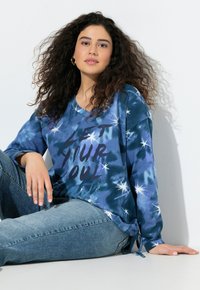 Blaues Batik-Sweatshirt mit V-Ausschnitt, das den Text "LASS DEINE SEELE" in fetten schwarzen Buchstaben und sternähnlichen Akzenten zeigt. Material aus Baumwollmischung.