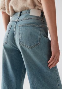 Lichtblauwe denim jeans met een losse pasvorm, voorzien van twee achterzakken en een witte leren patch op de achterkant van de tailleband. Zachte textuur.