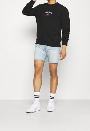 Personne portant un sweat-shirt noir Tommy Jeans, un short en denim bleu clair, des chaussettes blanches à rayures noires et des baskets blanches, debout contre un mur blanc.