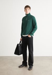 Grønn zip-up genser med høy krage, brukt over en hvit skjorte. Svart bukse, svart tote-bag og mørke sko fullfører antrekket.