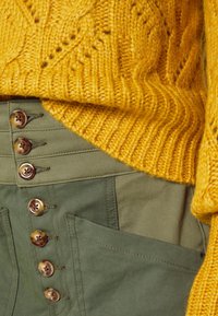 Geel gebreid sweater met geribbelde onderkant, laag over olijfgroene broek met hoge taille, voorzien van vijf ronde knopen en een zijzak.