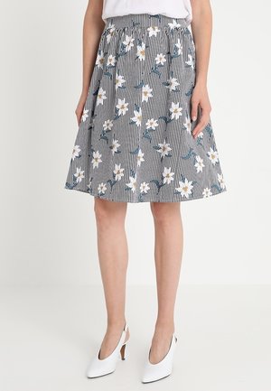Vrouw draagt een knielange rok met zwart-witte verticale strepen en wit bloemenpatroon, gecombineerd met witte slingback schoenen met hoge hakken.
