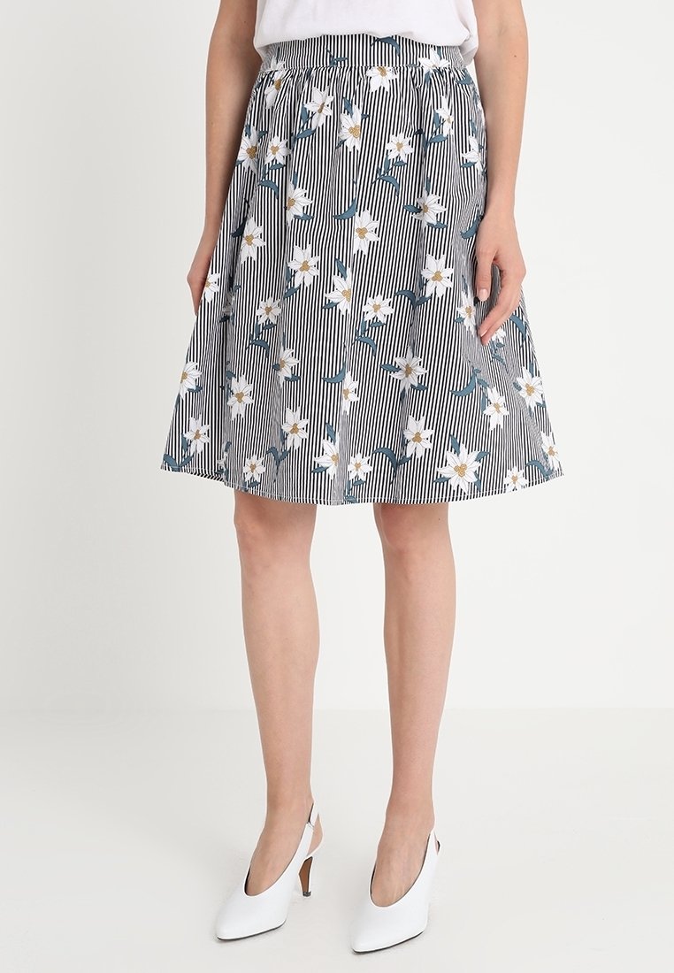Femme portant une jupe mi-longue à rayures verticales noires et blanches avec un motif de fleurs blanches, associée à des chaussures blanches à talons hauts et bride arrière.