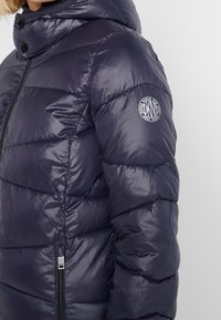 Giacca imbottita trapuntata blu scuro con cappuccio, zip nera e patch circolare del logo DKNY sulla manica superiore.