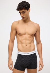 Melni bokseršorti ar ģeometrisku rakstu, Calvin Klein logos uz baltas jostas, elastīgs audums, pieguļošs dizains, bez redzamiem piederumiem.