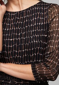 Vestido negro con estampado, con mangas transparentes, que presenta un diseño texturizado de formas alargadas en tonos de marrón y blanco. Detalles elegantes en los puños.