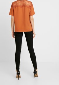 Blusa naranja óxido con mangas cortas y un panel de encaje en la espalda, combinada con pantalones ajustados negros y zapatos de tacón altos negros.