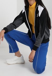 Svart och grå zip-up jacka med vita accenter, gul ribbad polokrage, blå sportbyxor med vita ränder och vita chunky sneakers.