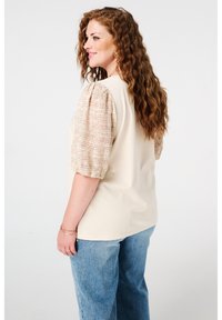Beige top met gestikte kanten pofmouwen, ontspannen pasvorm, gecombineerd met lichtblauwe jeans. De stof lijkt zacht met een gladde textuur.