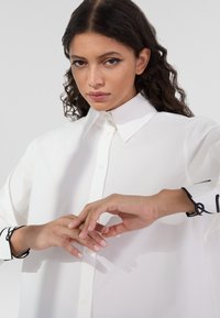 White oversized shirt made of smooth fabric, featuring a pointed collar and black accent embroidery on the cuffs. Model posing with hands.