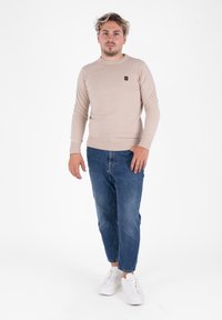 Suéter de punto acanalado beige con cuello redondo, combinado con jeans ajustados azules y zapatillas blancas. Incluye un pequeño logo en el suéter.